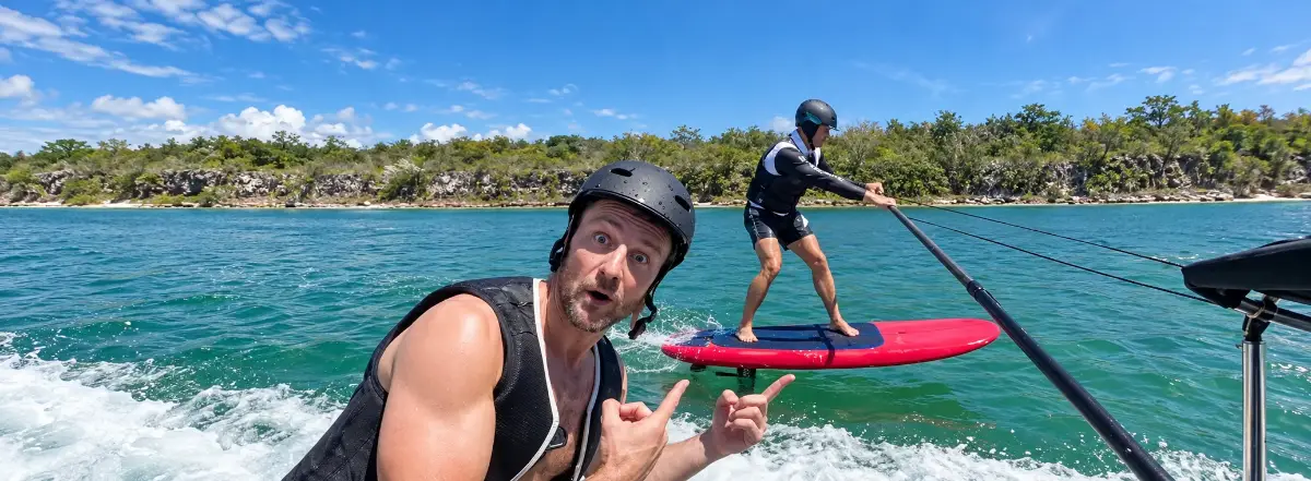 Rider on a red hydrofoil board being towed behind the boat while a student watches from the deck